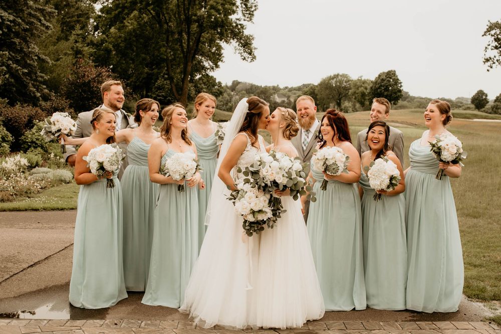 Bride and groom kissing surrounded by bridesmaids in sage green gowns holding white bouquets outdoors.