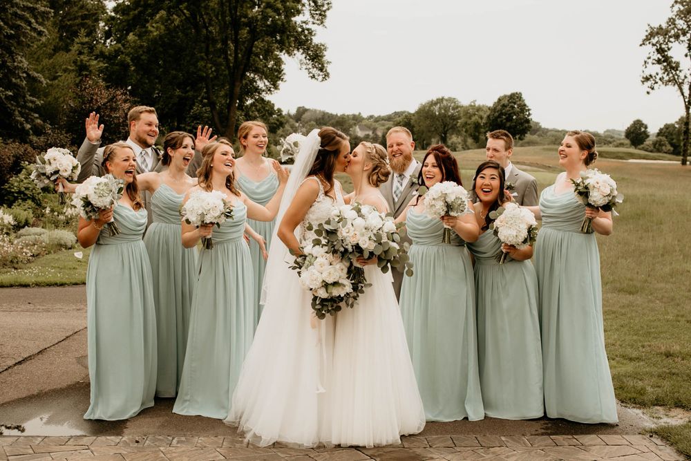 Two brides kiss surrounded by a joyful wedding party in mint green dresses and grey suits on a garden pathway.
