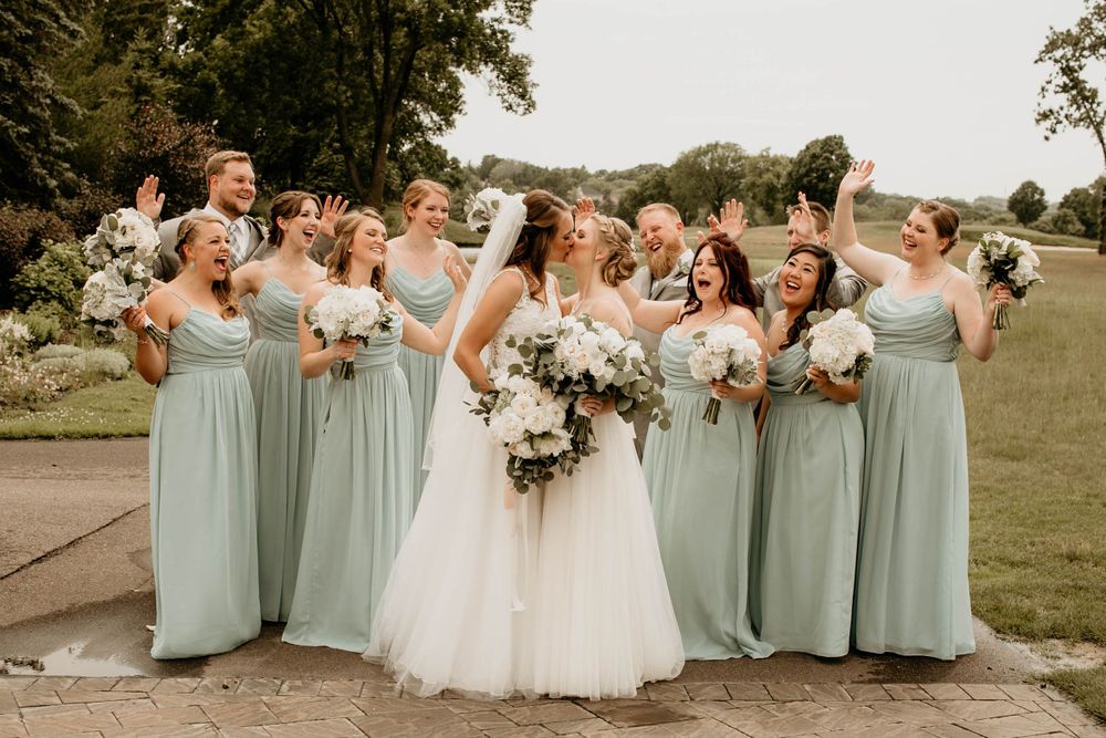 Joyful bridesmaids in mint green dresses celebrating with bride holding white floral bouquets outside.