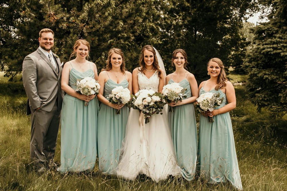 Bride in lace gown with bridesmaids and groomsman in sage green dresses posing in grassy field.
