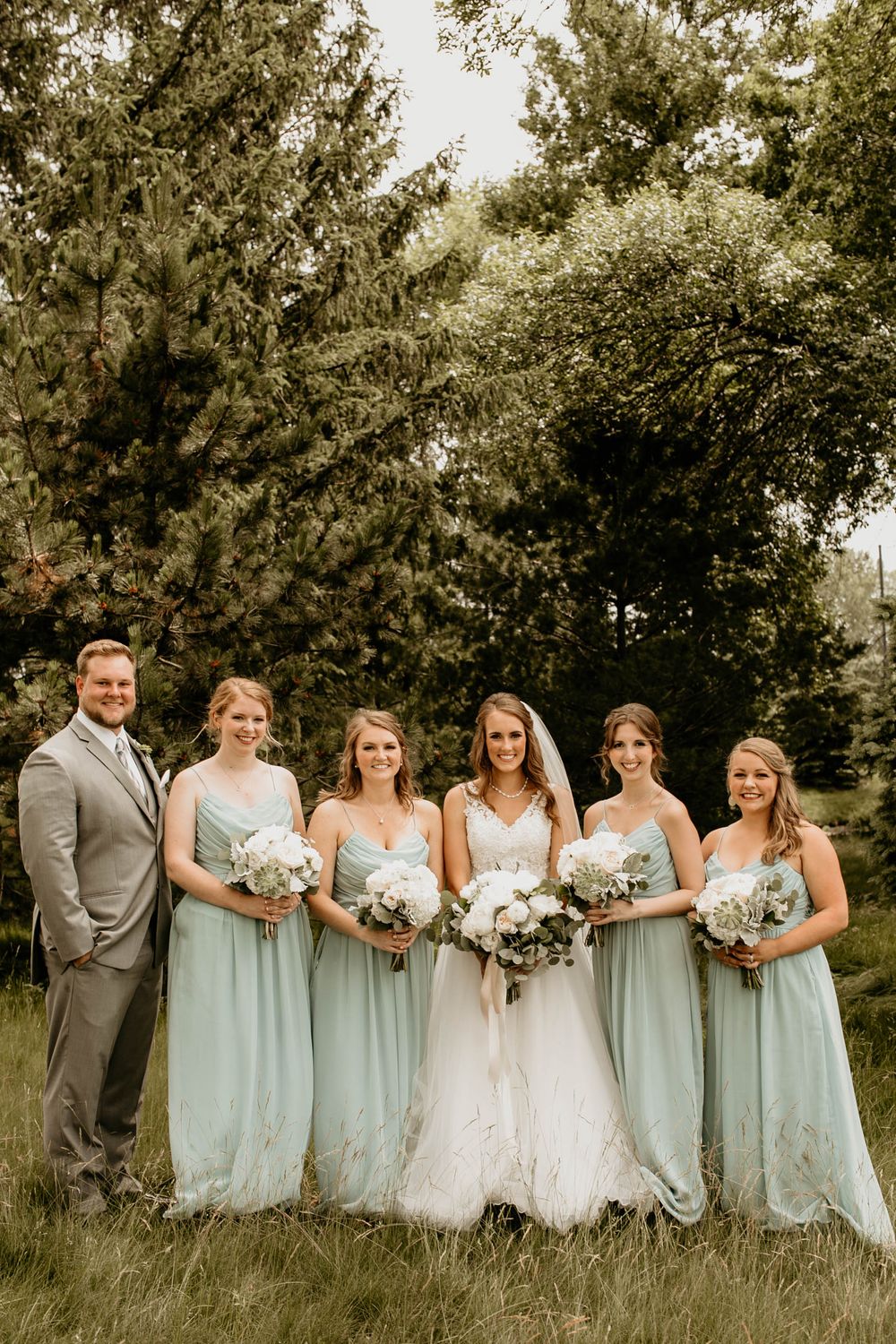 Bride with veil and bridesmaids in sage green gowns holding white bouquets before evergreen trees.