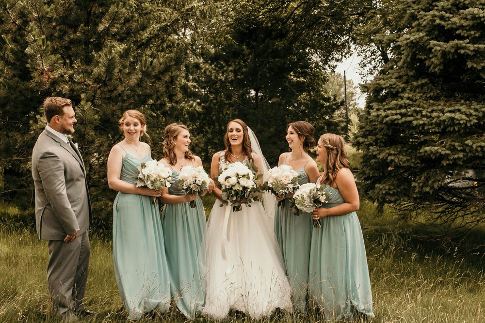 Bride and bridesmaids in mint dresses laughing together holding white bouquets in a green setting.