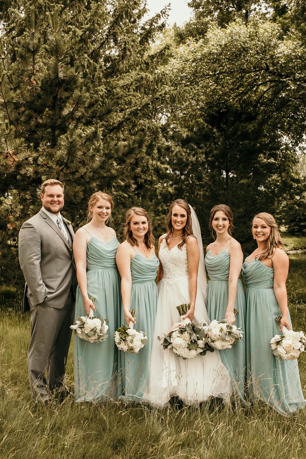 Wedding party posing in field with bridesmaids in mint gowns holding white bouquets by tall trees.