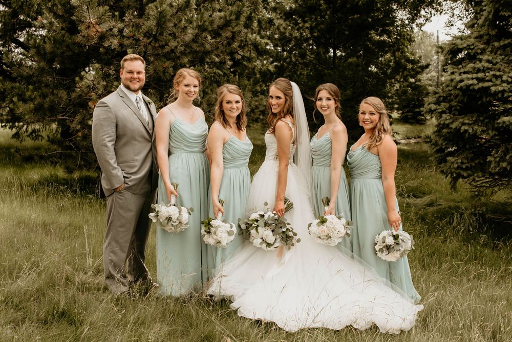 Smiling wedding party with bridesmaids in sage green dresses and groomsman posing in grassy field.