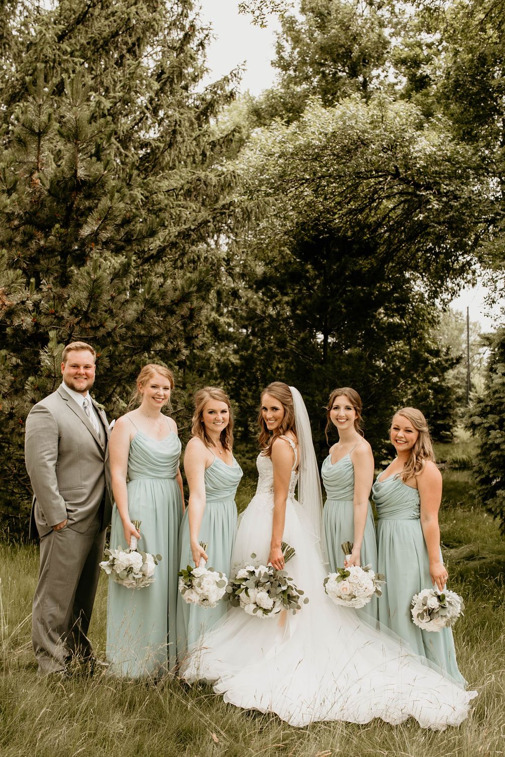 Bride in white gown with veil and bridesmaids in sage green dresses posing in lush green field.