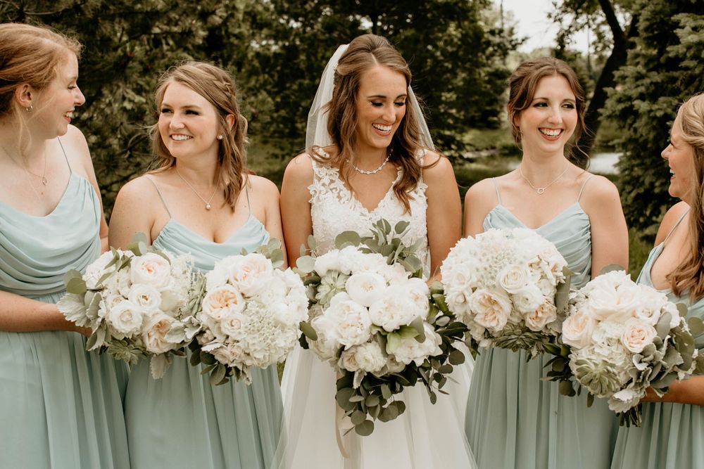 Bride in white lace gown smiles with bridesmaids in sage green dresses holding white floral bouquets outdoors.