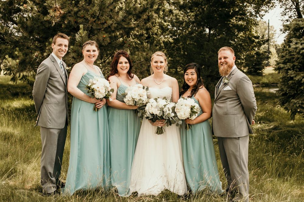 Wedding party poses outdoors with bride in white gown, bridesmaids in sage dresses, and groomsmen in gray suits.