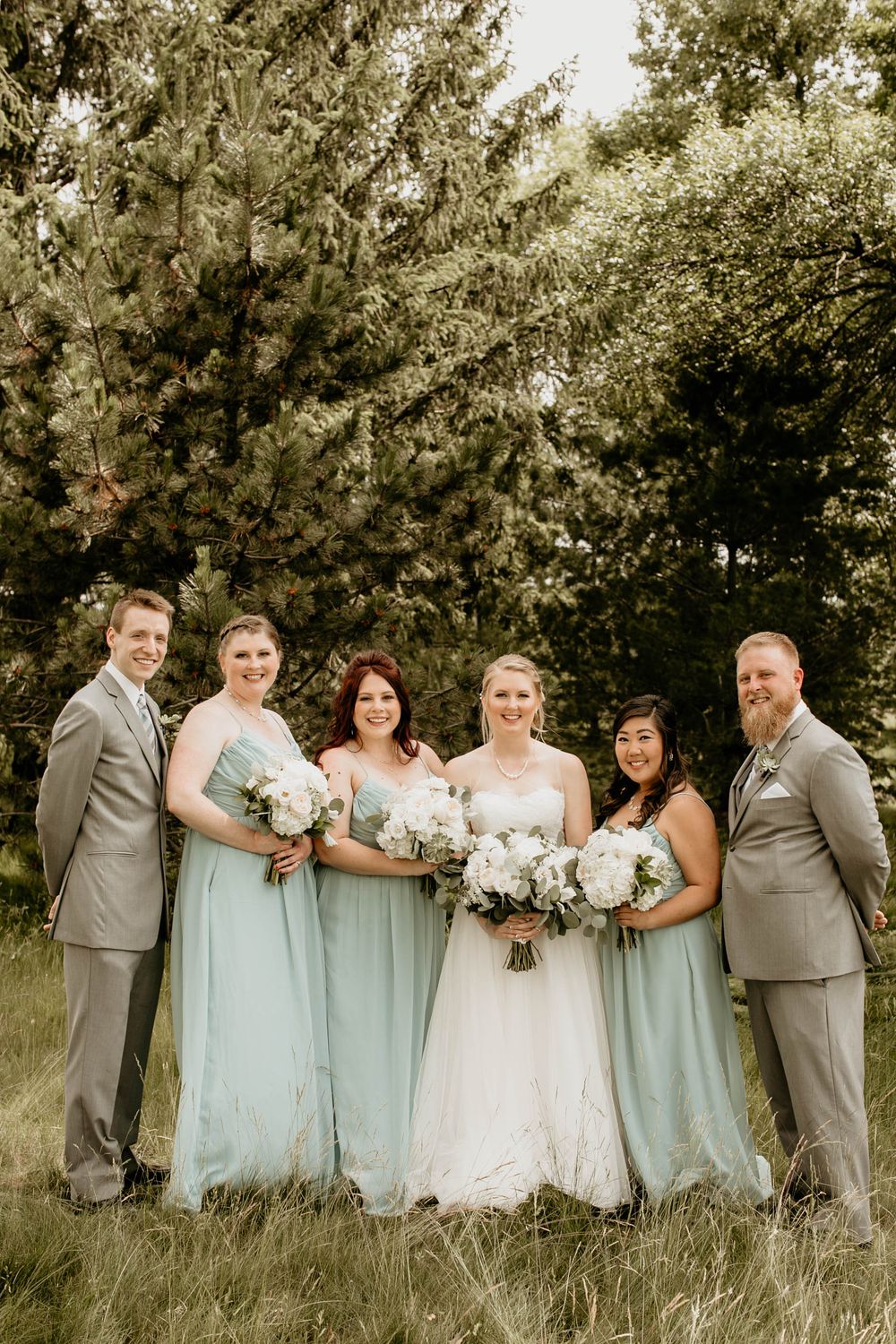Wedding party of six smiles in a grassy field, bridesmaids in mint gowns and groomsmen in gray suits.