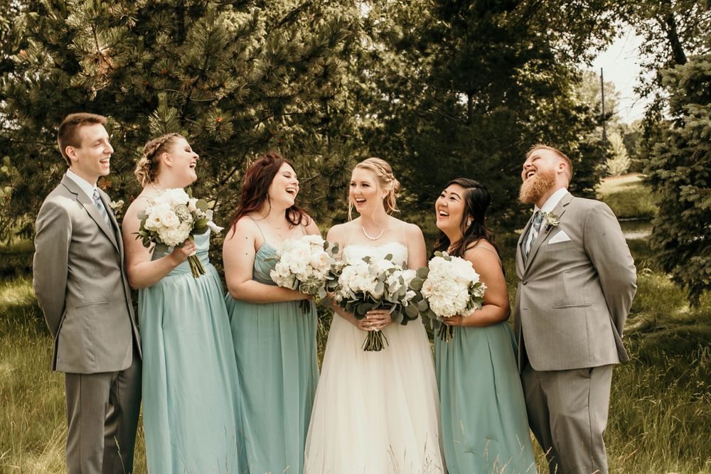 Bride and wedding party laugh together outdoors in green field, wearing sage bridesmaid dresses and gray suits.