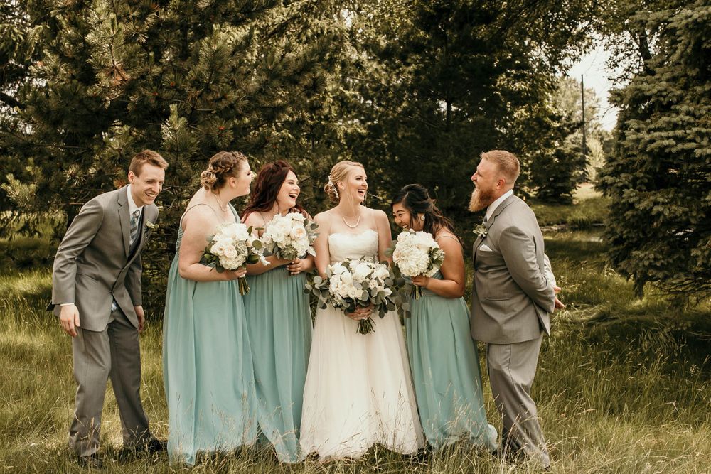 Joyful wedding party laughs together in a lush green field, bridesmaids in sage dresses with white bouquets.