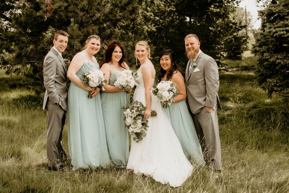 Smiling wedding party poses in sunlit grassy field, bridesmaids in mint gowns and groomsmen in gray suits.