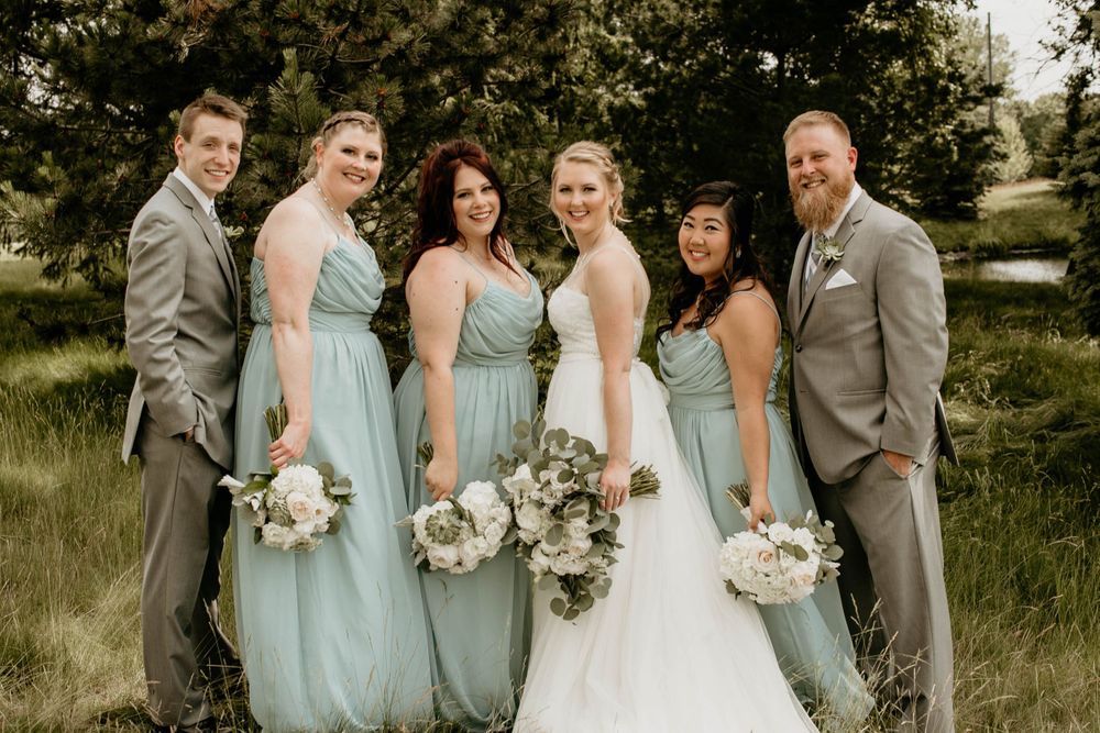 Wedding party of six smiles in green field, bridesmaids in sage dresses holding white bouquets with groomsmen.