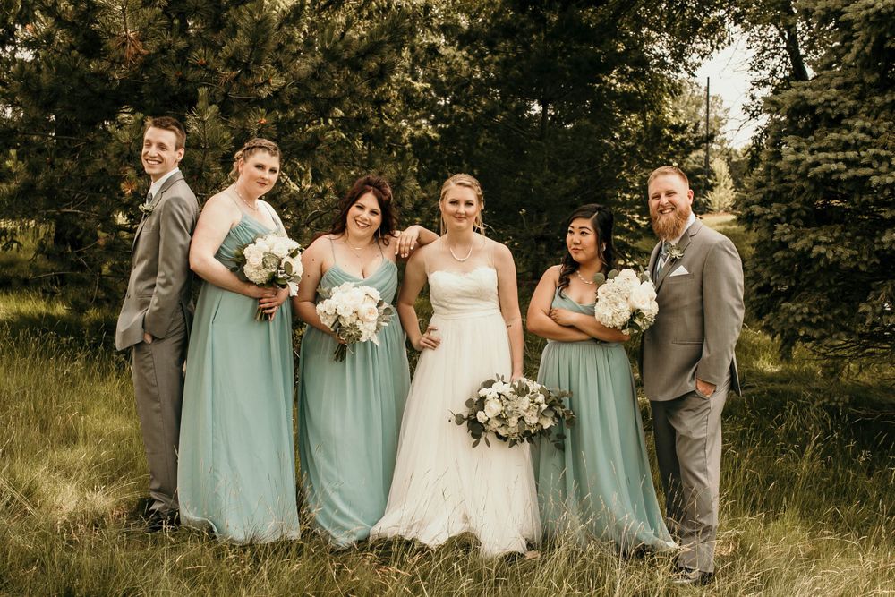 Wedding party poses casually in green meadow, bridesmaids in sage dresses and groomsmen in gray suits outdoors.