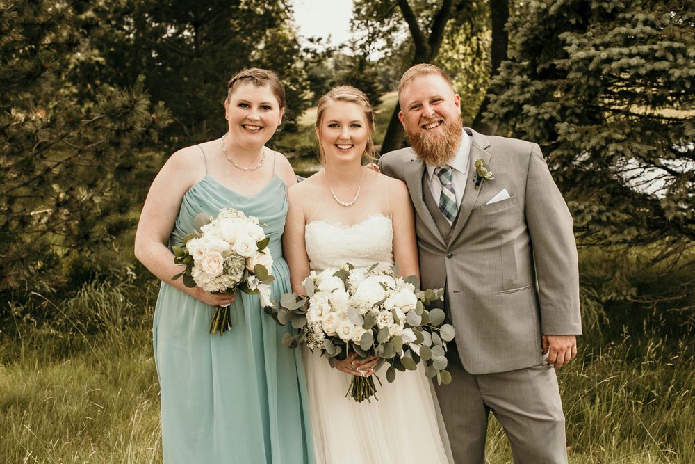 Bride in white strapless gown poses with bridesmaid and groomsman holding white bouquets in a green field.