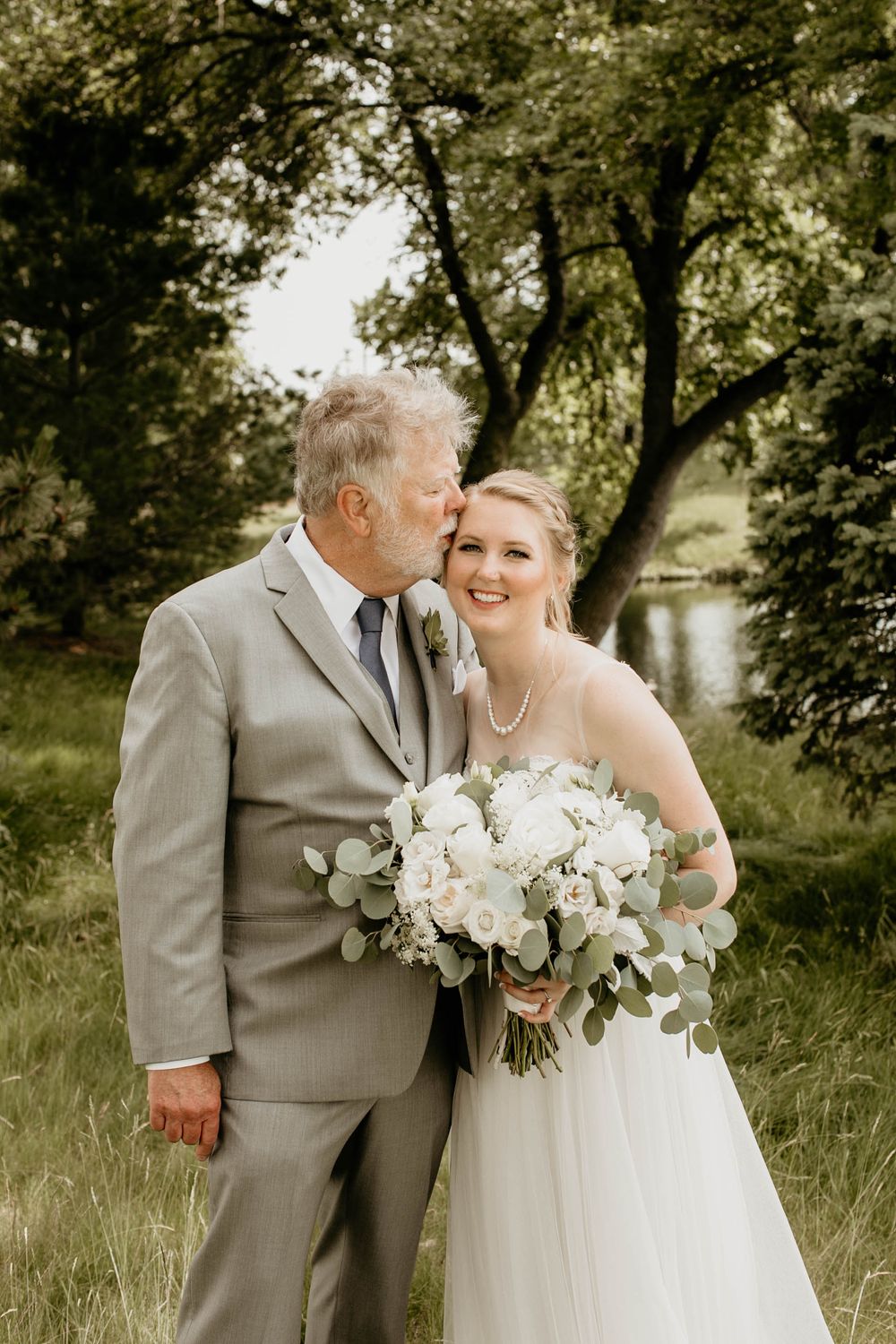 Older gentleman in gray suit kisses bride on cheek as she holds white rose bouquet near a pond.