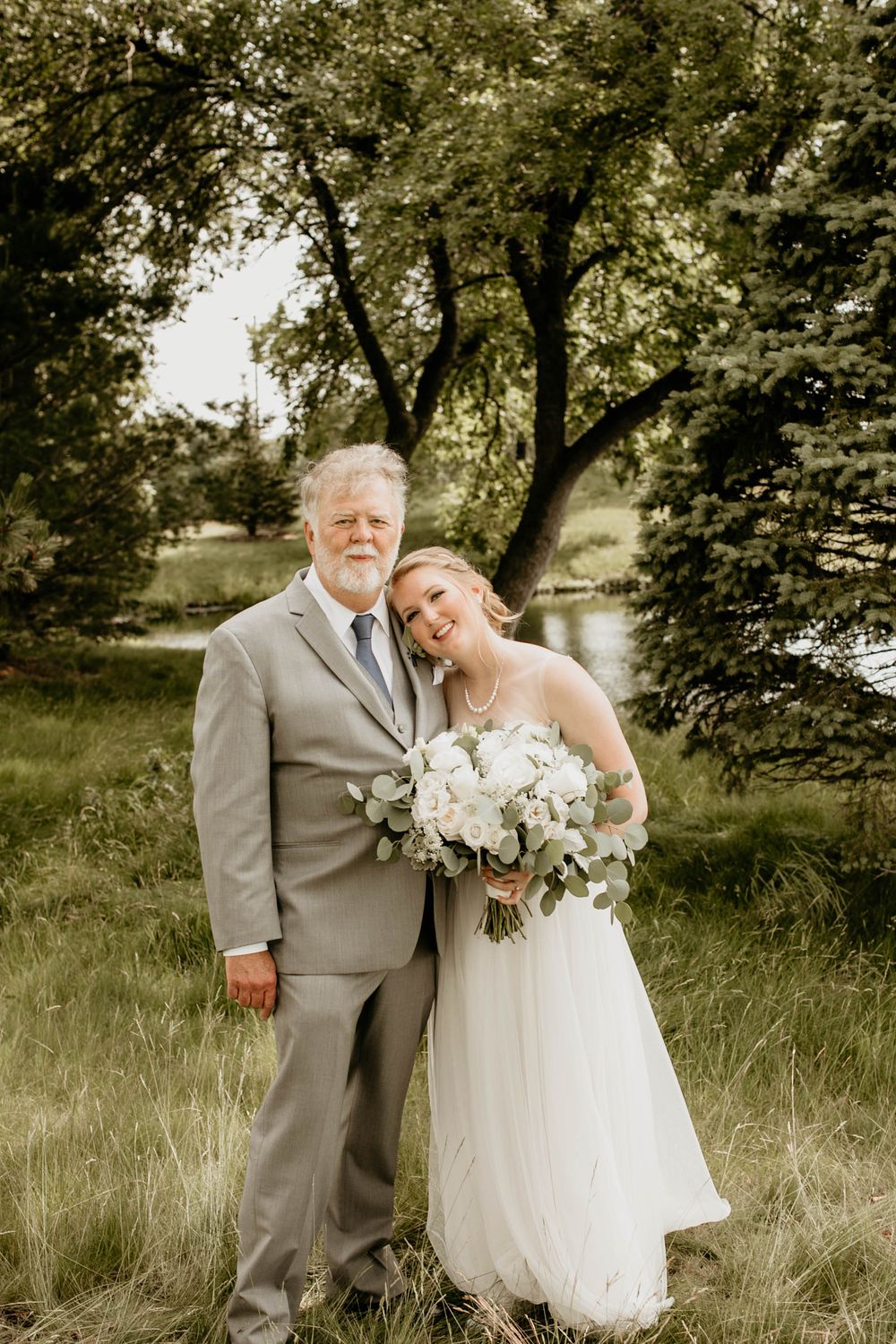 Bride in white gown holds bouquet beside older man in gray suit outdoors at wedding.