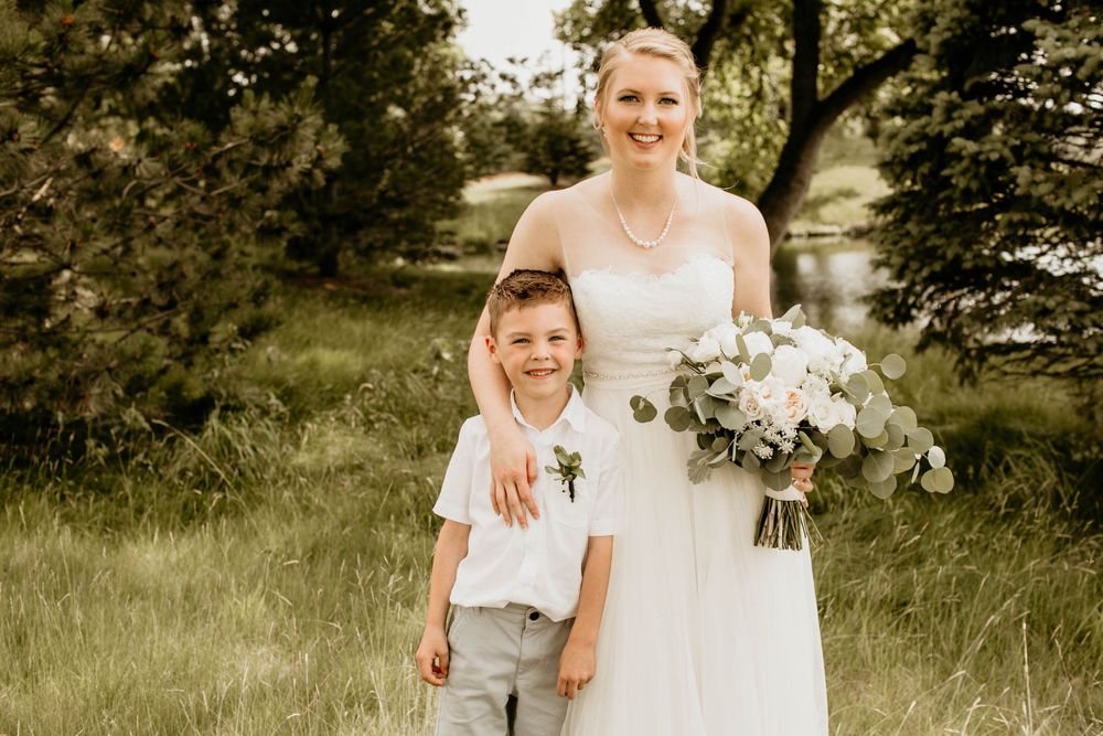 Smiling bride in strapless white gown poses with young boy in white shirt at outdoor wedding.