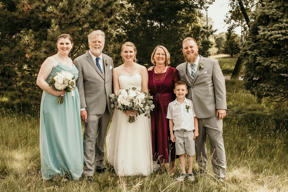Wedding party of six poses outdoors including bride in white gown and family in formal attire.