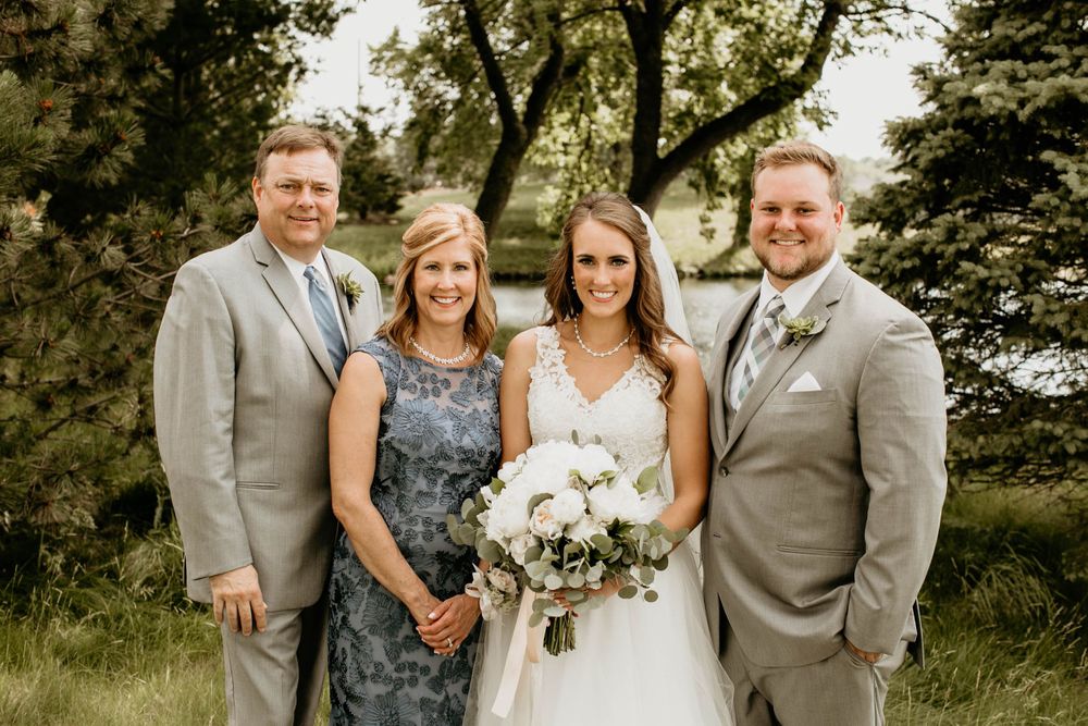 Bride with white bouquet poses with three family members in gray suits and blue dress outdoors.
