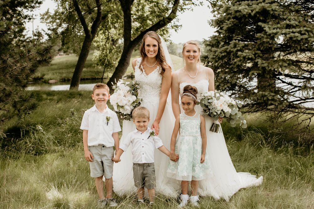 Two brides in white gowns pose with three young children in wedding attire on grassy lawn.