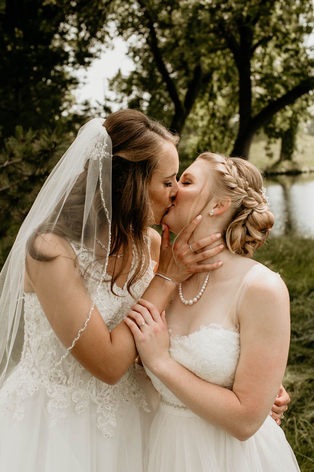 Two brides in white wedding gowns share a romantic kiss outdoors near trees and water.