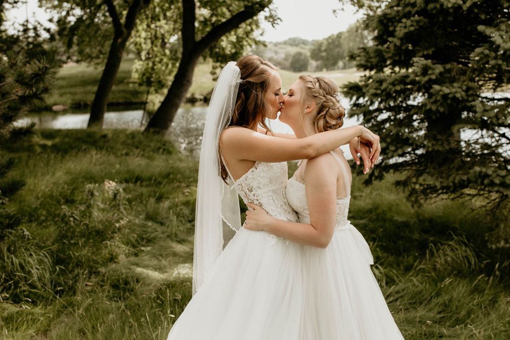 Two brides in white gowns embrace and kiss in a lush green outdoor setting.