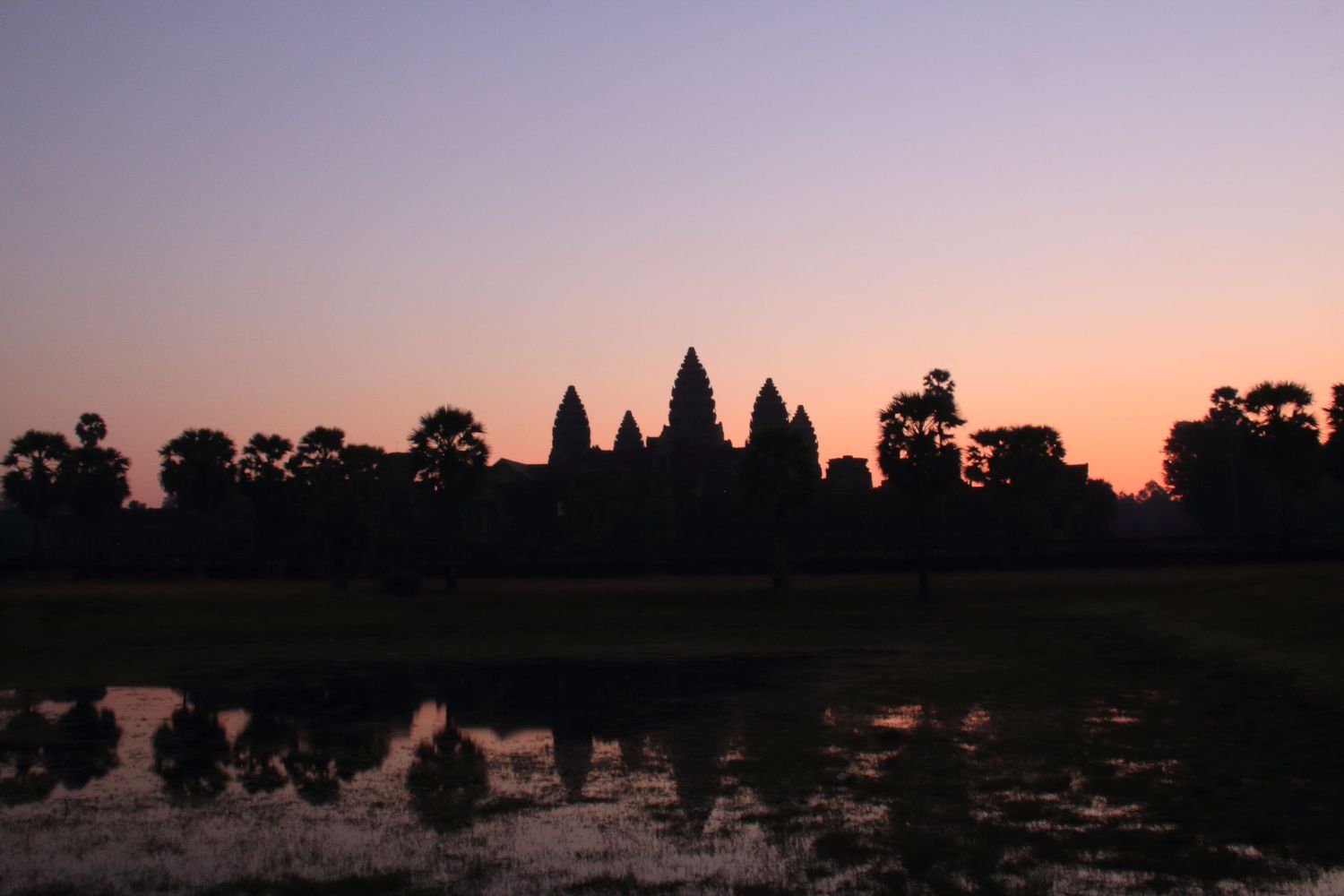 Silhouette of Angkor Wat temple spires reflected in water during sunset.