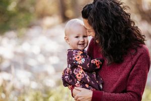 Curly-haired mother tenderly smiling at her happy baby girl in a floral outfit in a sunlit outdoor setting.