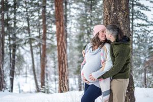 A couple embraces by a snow-covered pine tree in a winter forest, cradling a pregnant belly.