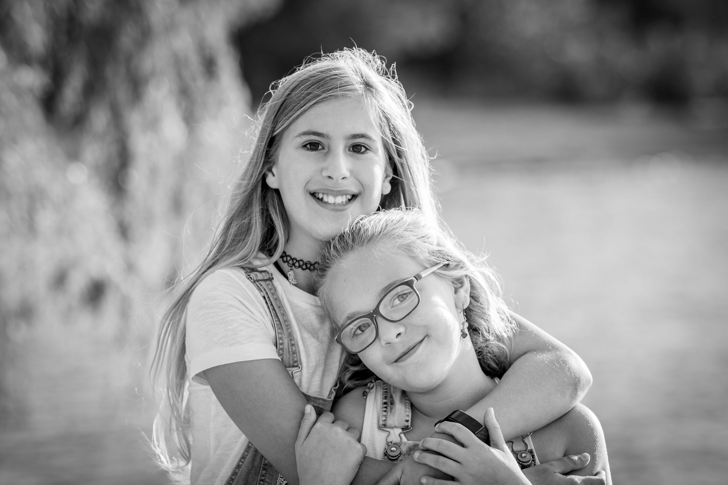 Black and white portrait of two smiling sisters hugging closely outdoors in soft natural light.