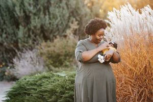 A woman in a gray dress tenderly holds her newborn baby among tall ornamental grasses outdoors.