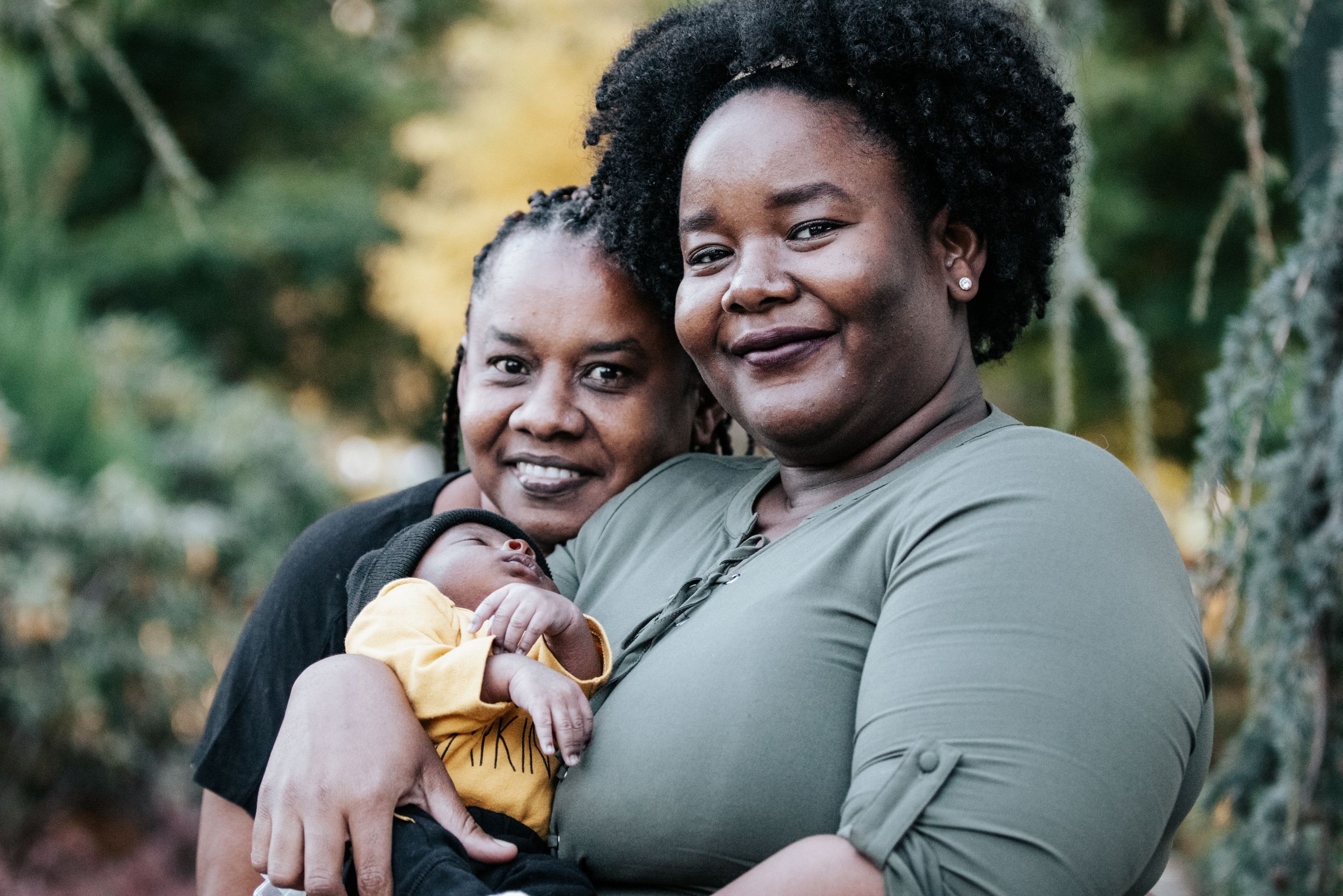 Two smiling women pose together outdoors, one holding a newborn baby wrapped in yellow fabric.
