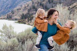 A laughing mother carries two young children in rust outfits on a scenic mountain lakeside trail.