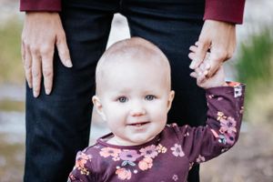Adorable smiling baby girl in a floral top standing and holding an adult's hand outdoors in natural light.
