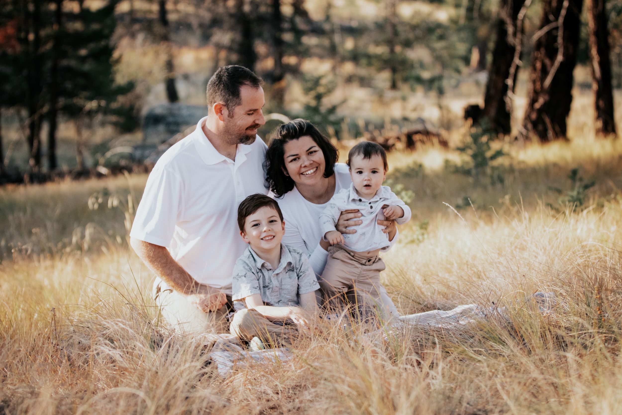 Happy family of four sits in golden grass field, parents holding two young children outdoors.