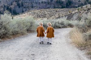 Two toddlers in matching rust-colored dresses hold hands walking down a rural dirt path together.