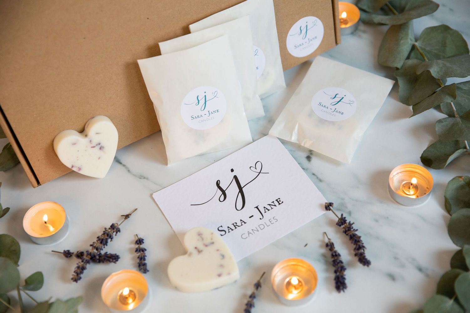White gift packages with monogram logo displayed with lit candles, eucalyptus leaves and decorative heart accents from a product photoshoot.