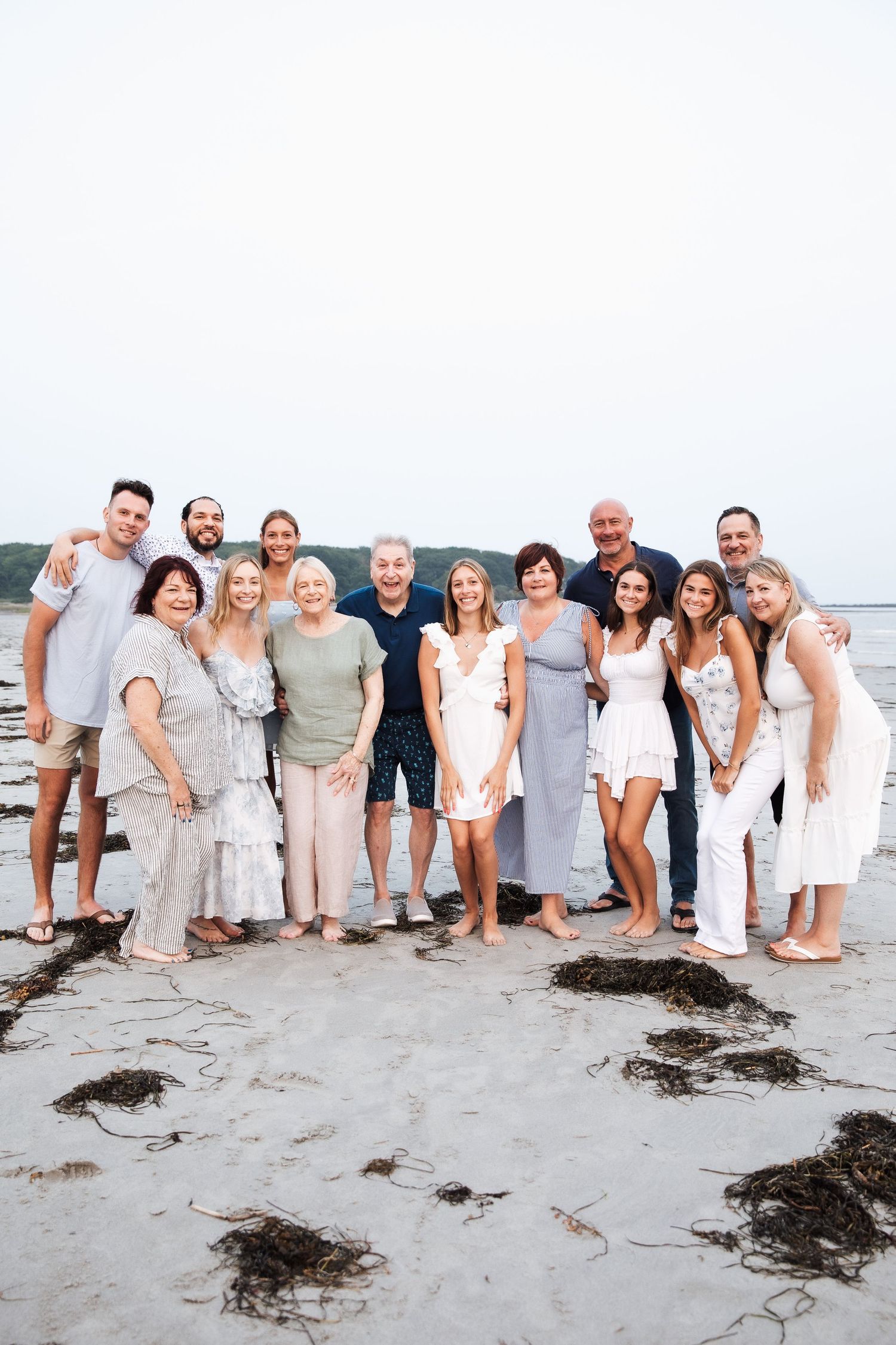 Large group in coordinated white and neutral outfits posing together on beach at sunset. extended family photoshoot