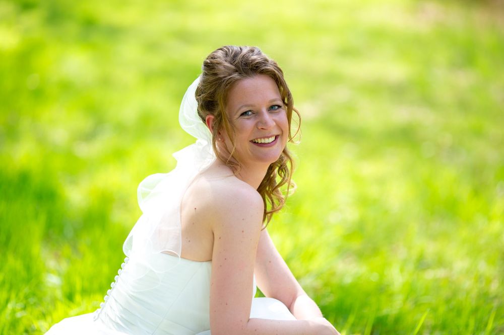 Bride in white dress smiling while sitting in a sunlit meadow.