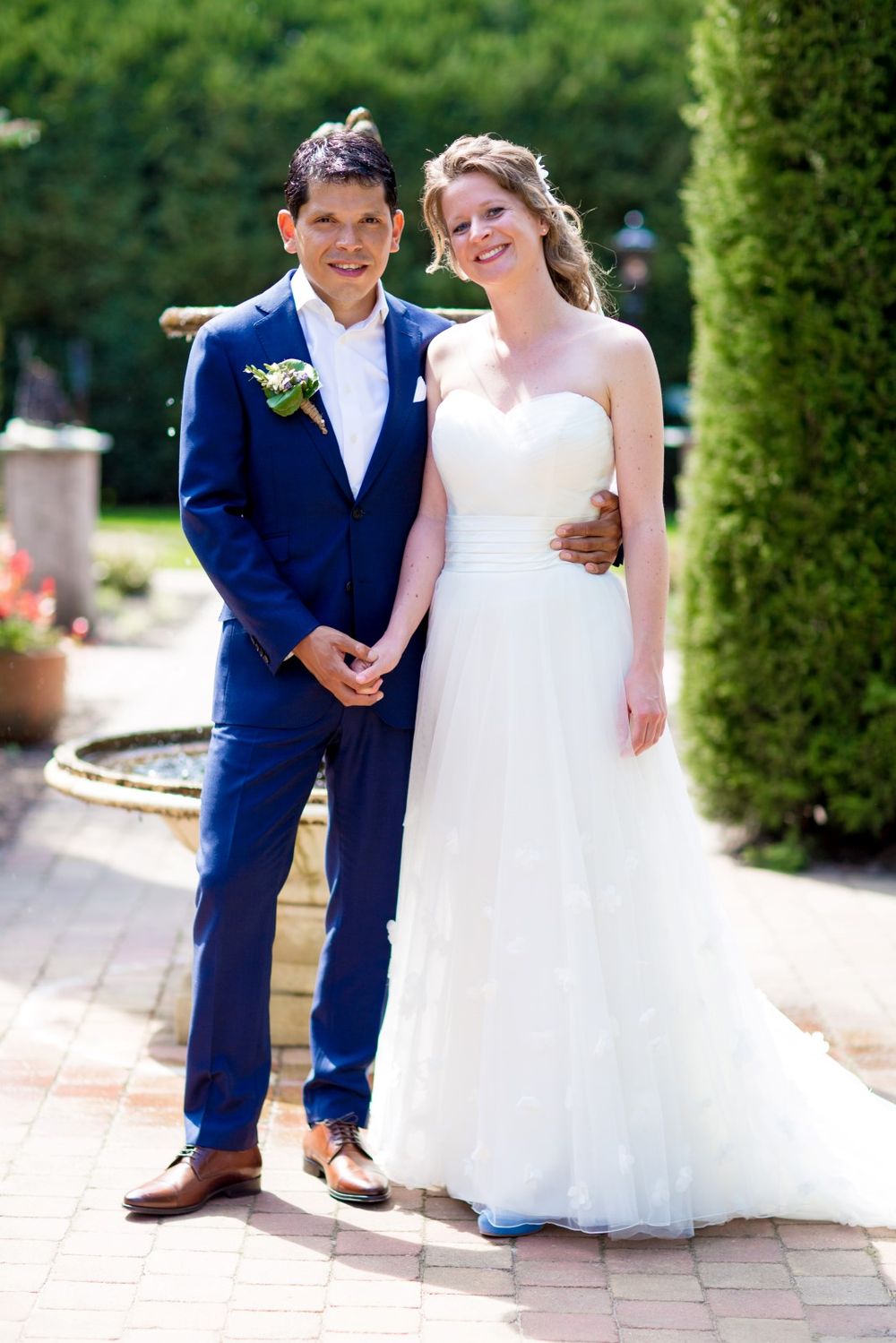 Elegant wedding couple standing by a garden fountain in formal attire.