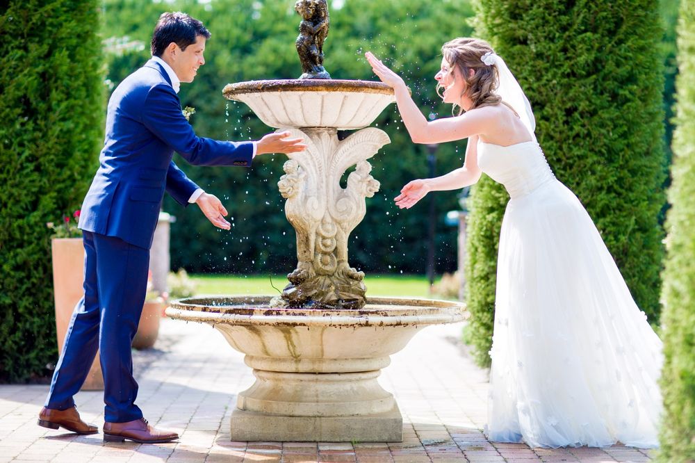 Playful wedding couple splashing water at each other by a garden fountain.