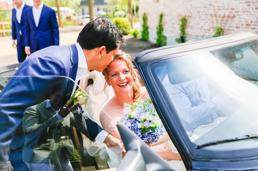 A wedding couple shares an intimate moment while getting into a car with blue flowers in the bouquet.