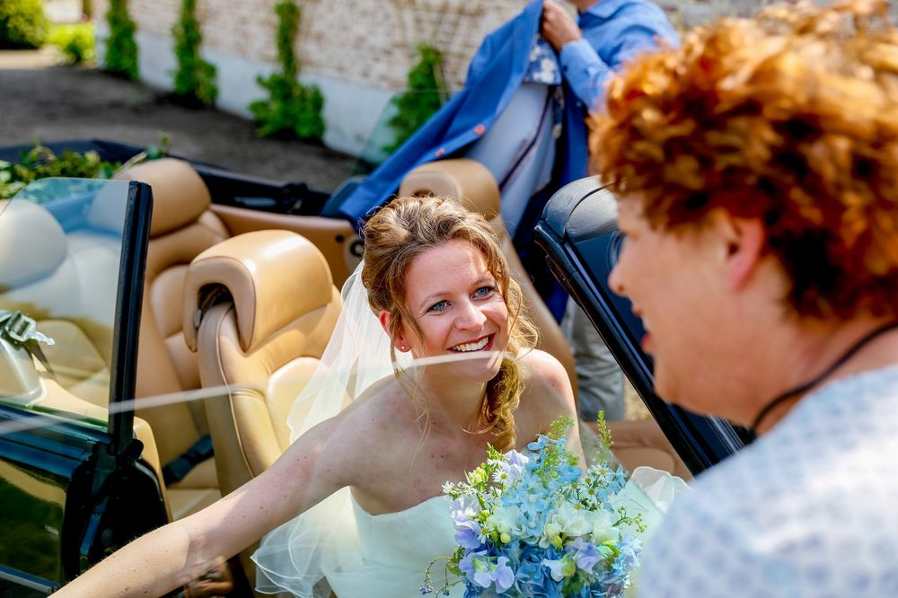 A bride with a bouquet sits in a convertible car during a wedding celebration.