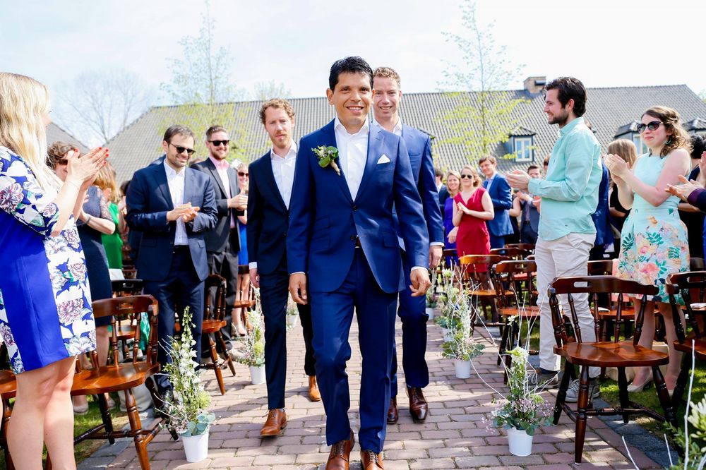 A groom in a navy suit walks down an outdoor aisle lined with wooden chairs and floral decorations.