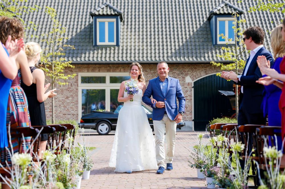Wedding guests watch as a couple walks through a flower-lined path towards a stone building.