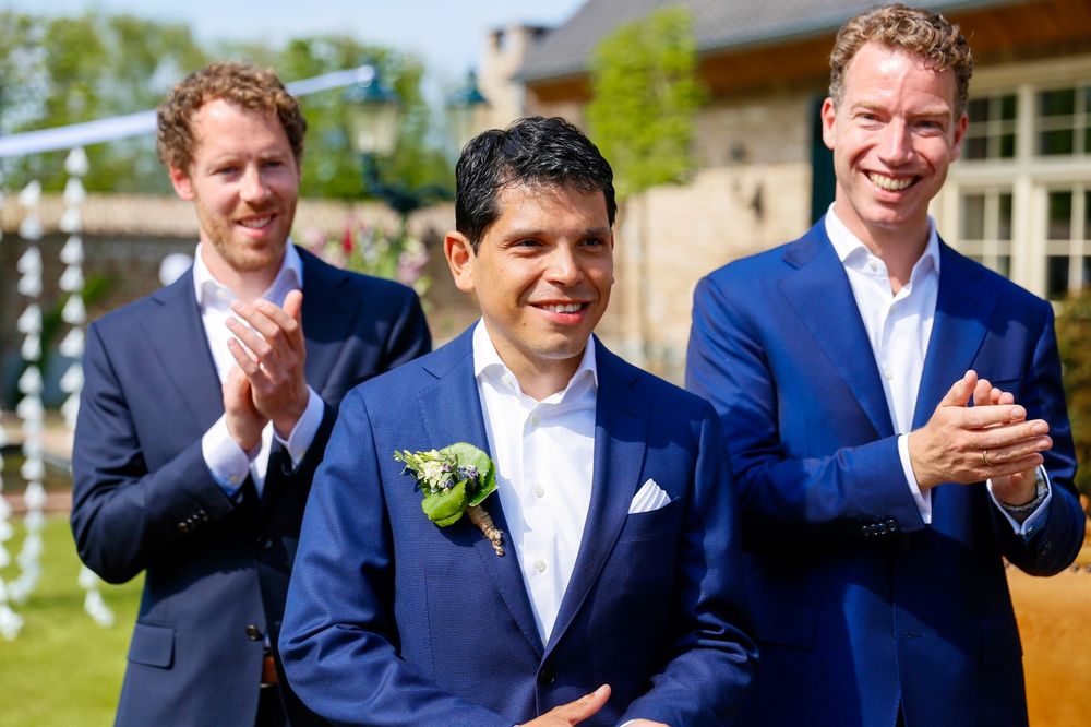 Groomsmen in blue suits applaud during an outdoor wedding ceremony.