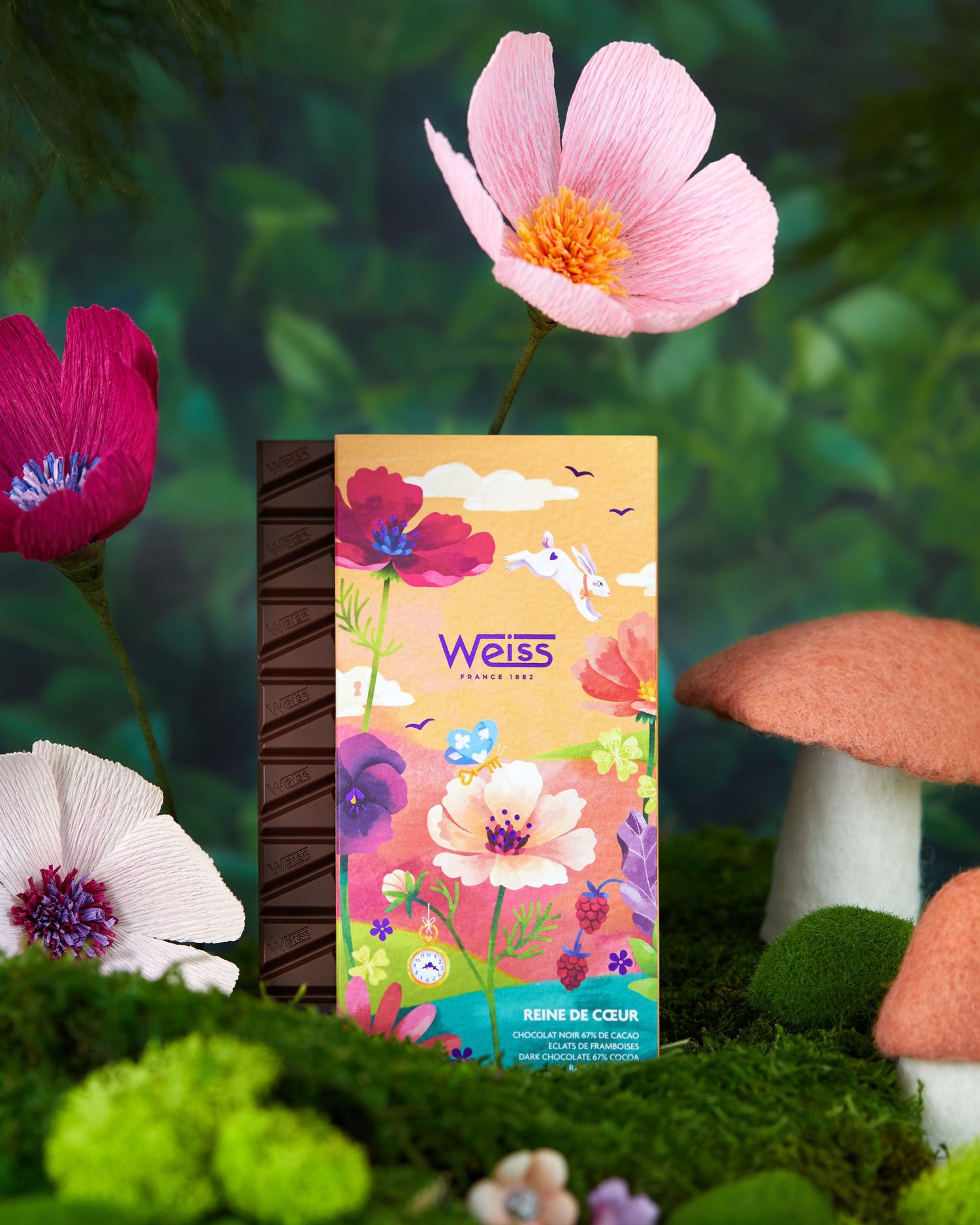 A colorful Weiss chocolate package with floral artwork sits among pink flowers and mushrooms on mossy ground.