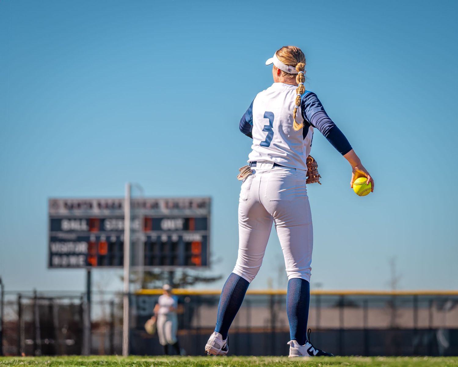 Softball player in white uniform holding yellow ball against blue sky with scoreboard in background.