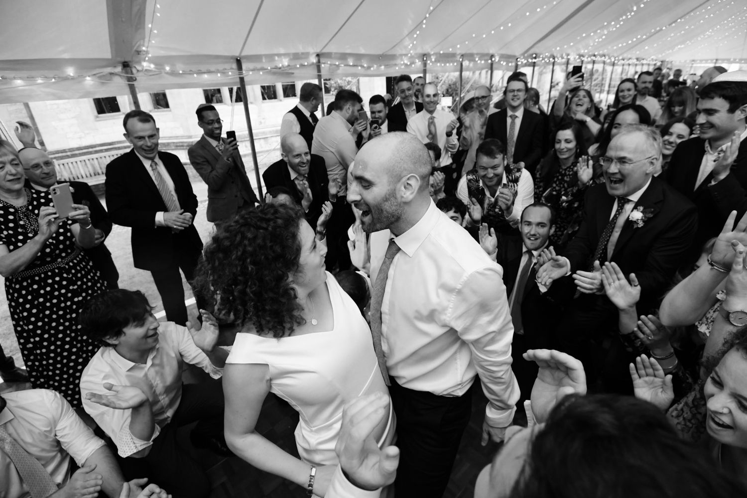 Wedding guests dancing and celebrating under white tent during reception party.