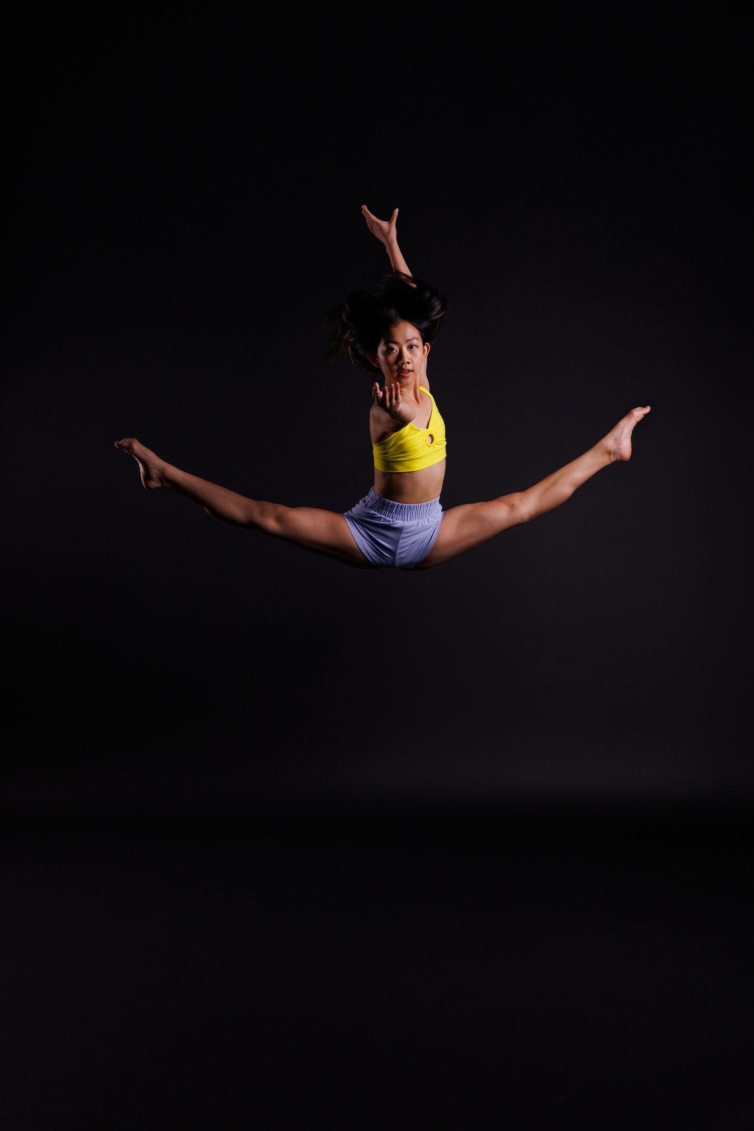 Dancer executes perfect split jump in yellow top and shorts against dark background.